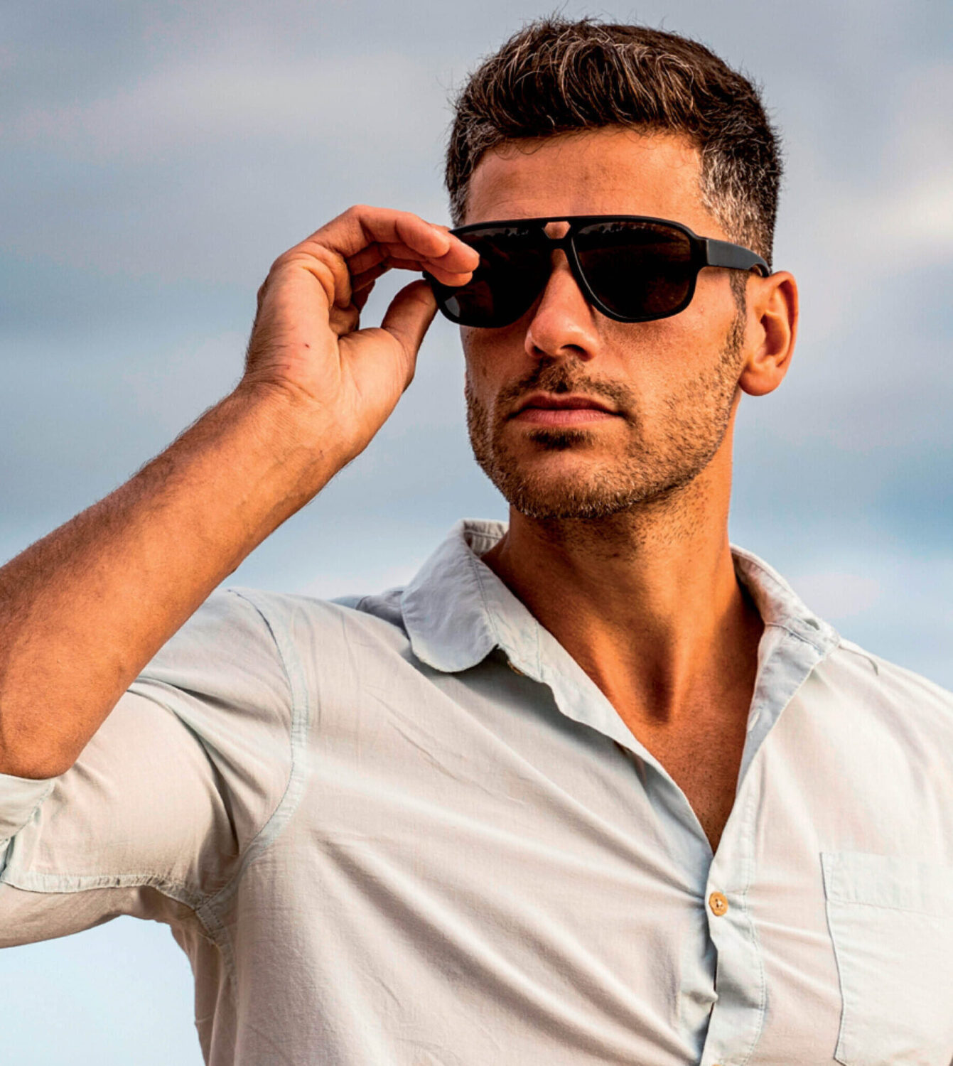 Een man met een stoppelbaard draagt stijlvolle zwarte zonnebrillen en een lichtblauw overhemd in de buitenlucht.